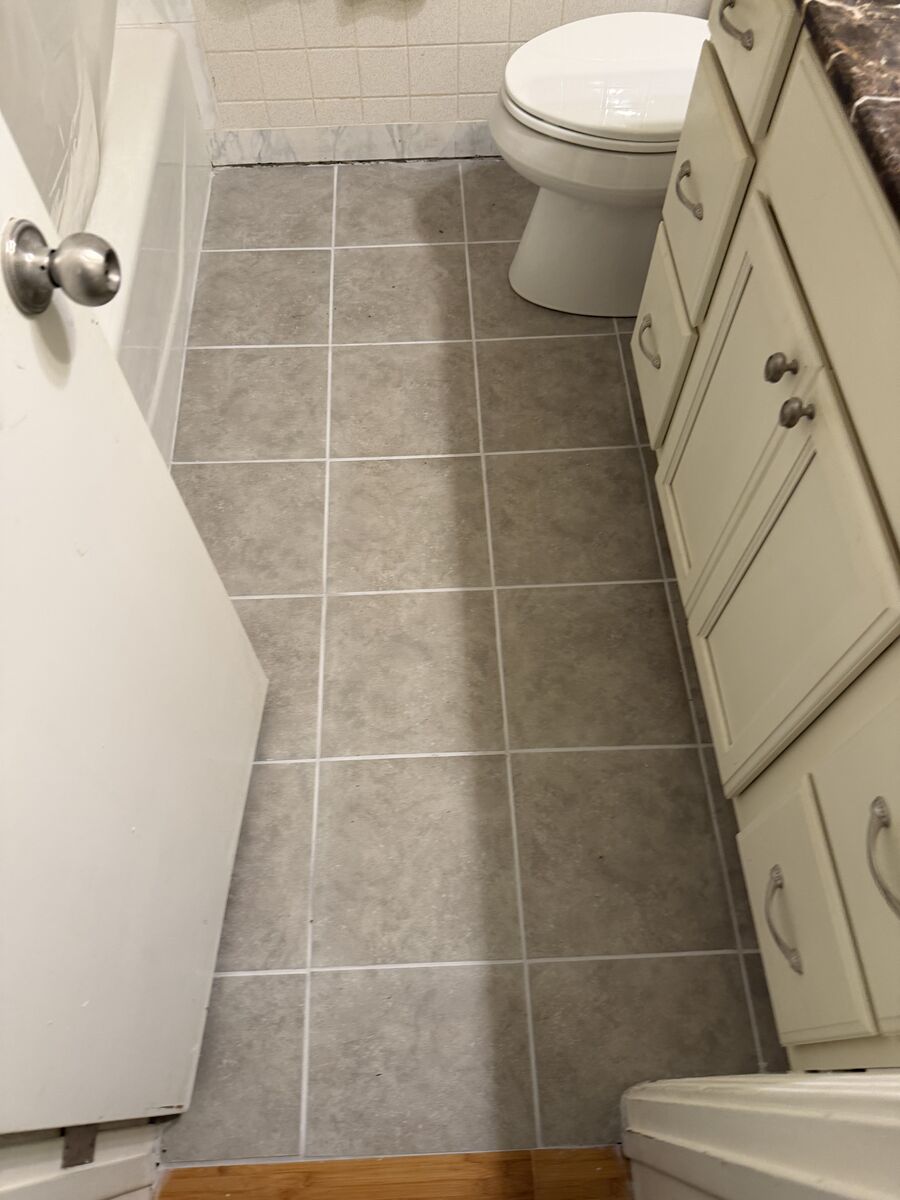 Newly installed gray tile floor in a narrow bathroom, seen from the doorway, with white grout lines running between the vanity and tub